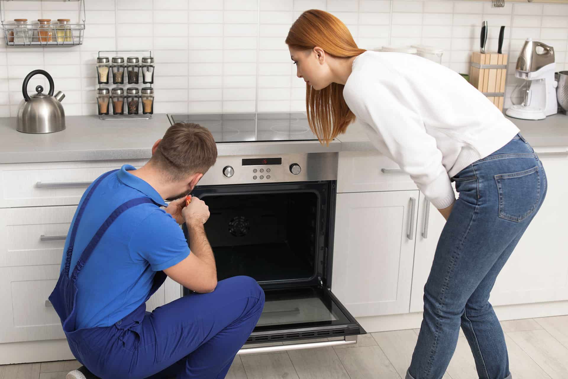 A male mechanic fixing an oven with woman watching him